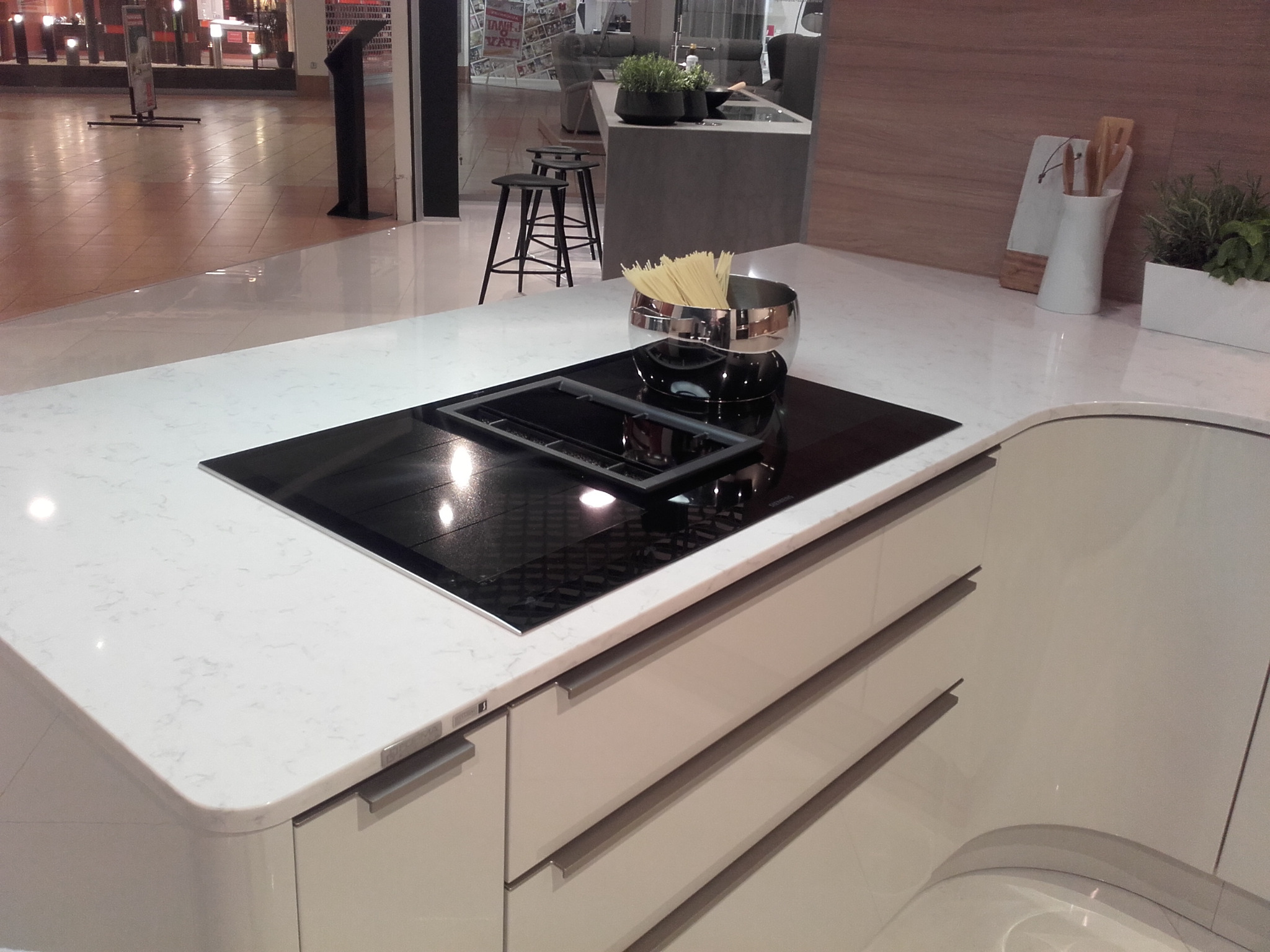 Biały blat kuchenny z marmurowym wzorem, wbudowana czarna płyta indukcyjna, metalowa misa z makaronem, w tle widoczny fragment wystawy meblowej.