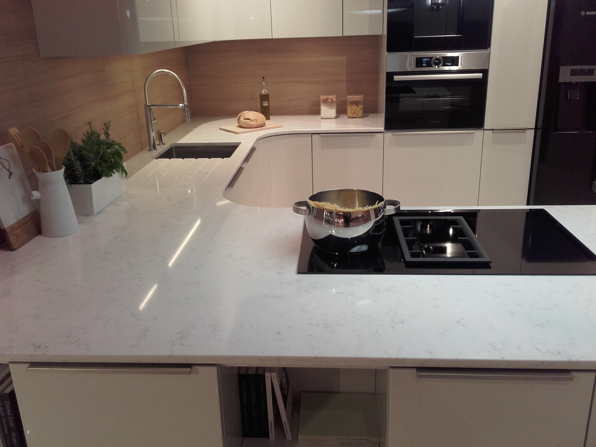 Jasny blat kuchenny z kamienia kompozytowego, z wbudowaną płytą grzewczą i garnkiem z gotującym się makaronem, nowoczesna kuchnia z drewnianym panelem ściennym i wbudowanymi sprzętami AGD.