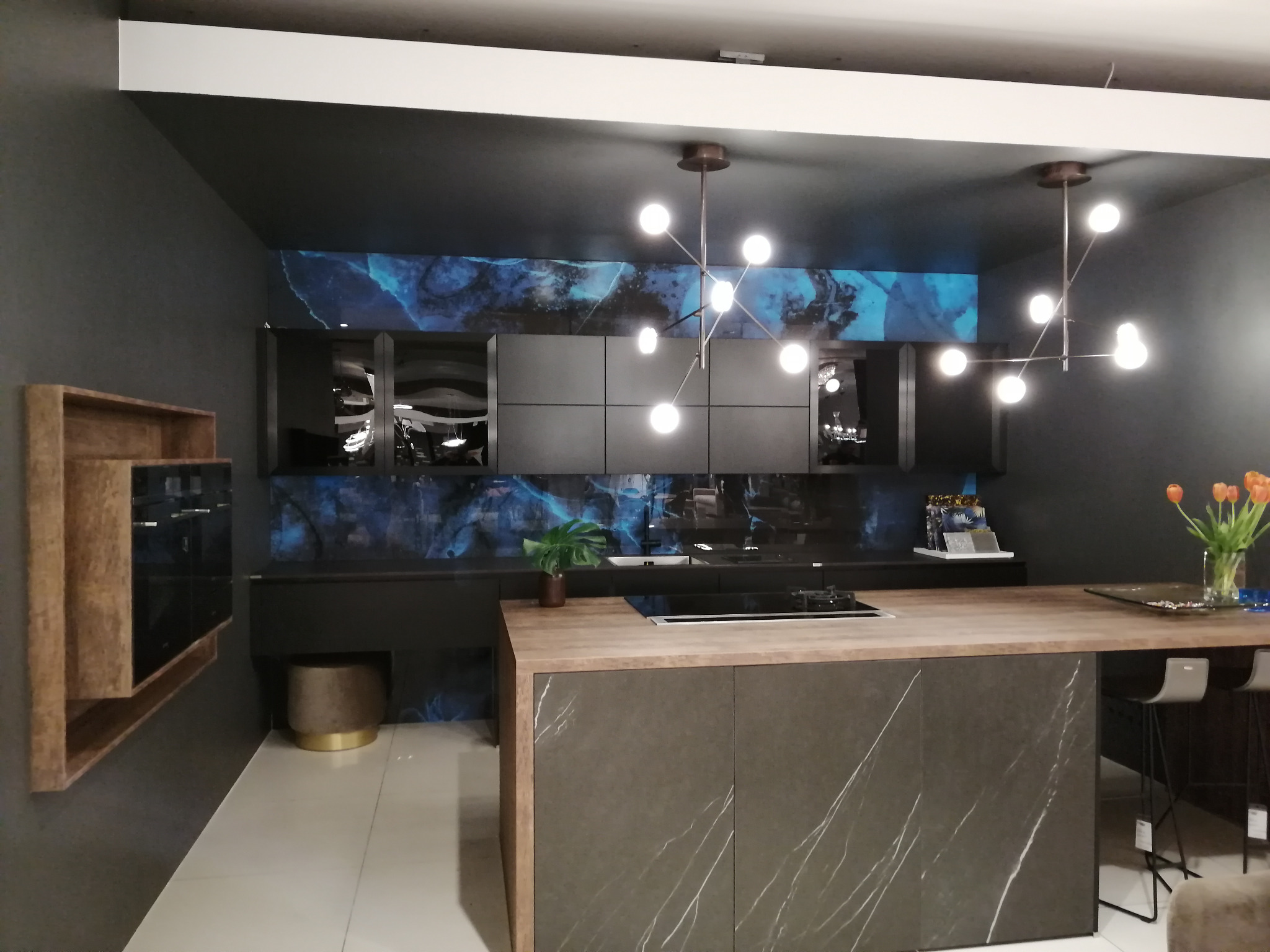 Wnętrze nowoczesnej kuchni z wyspą wykończoną ciemnym kamieniem z jasnymi żyłami, drewniany blat, czarne szafki wiszące na tle ściany z motywem marmuru, nowoczesne oświetlenie.