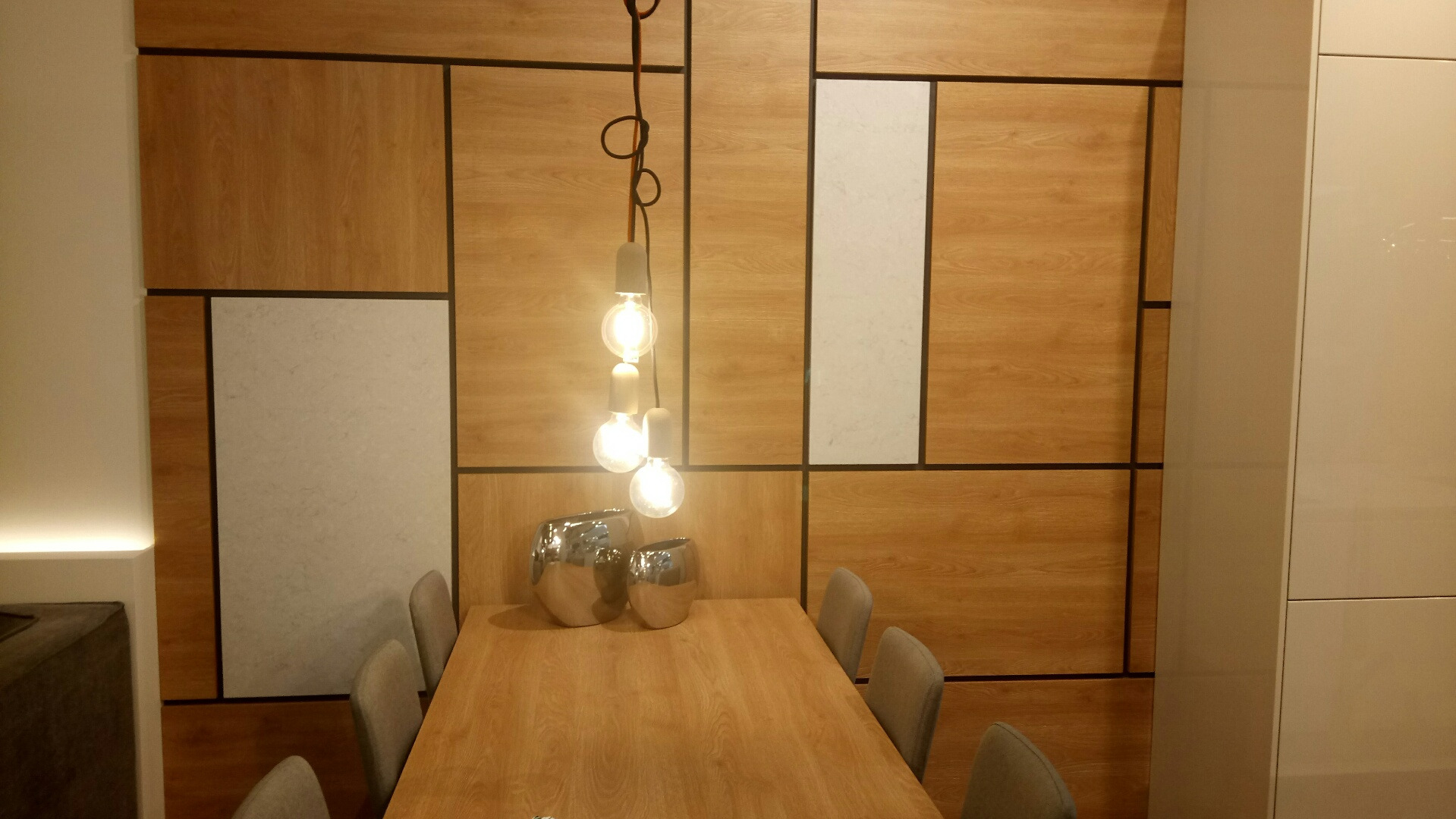 Jadalnia z drewnianym stołem, szarymi krzesłami i dekoracyjną ścianą z drewnianych i kamiennych paneli w geometryczny wzór, oświetlona wiszącą lampą z trzema żarówkami i ozdobiona dwoma srebrnymi...