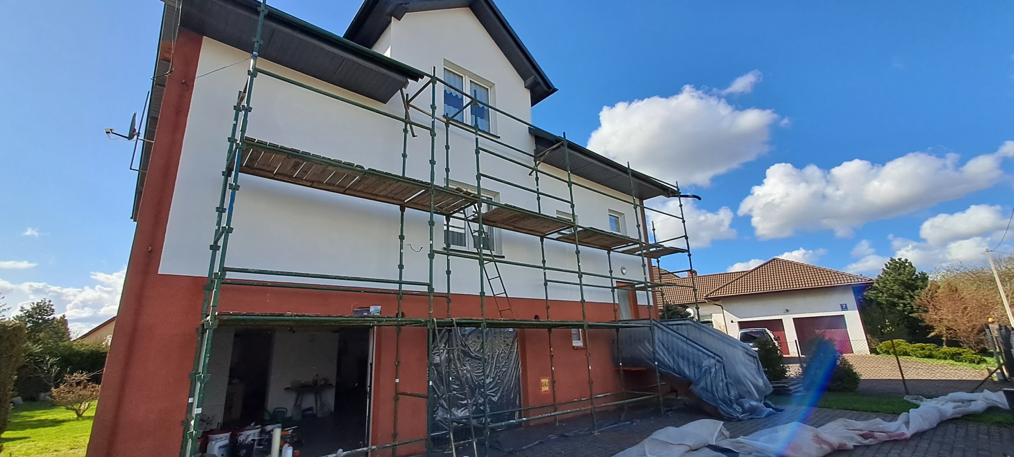 Dom jednorodzinny w trakcie malowania elewacji, widoczne rusztowanie, fragment odsłoniętej, pomarańczowej ściany i biała, świeżo pomalowana część, błękitne niebo z chmurami w tle.