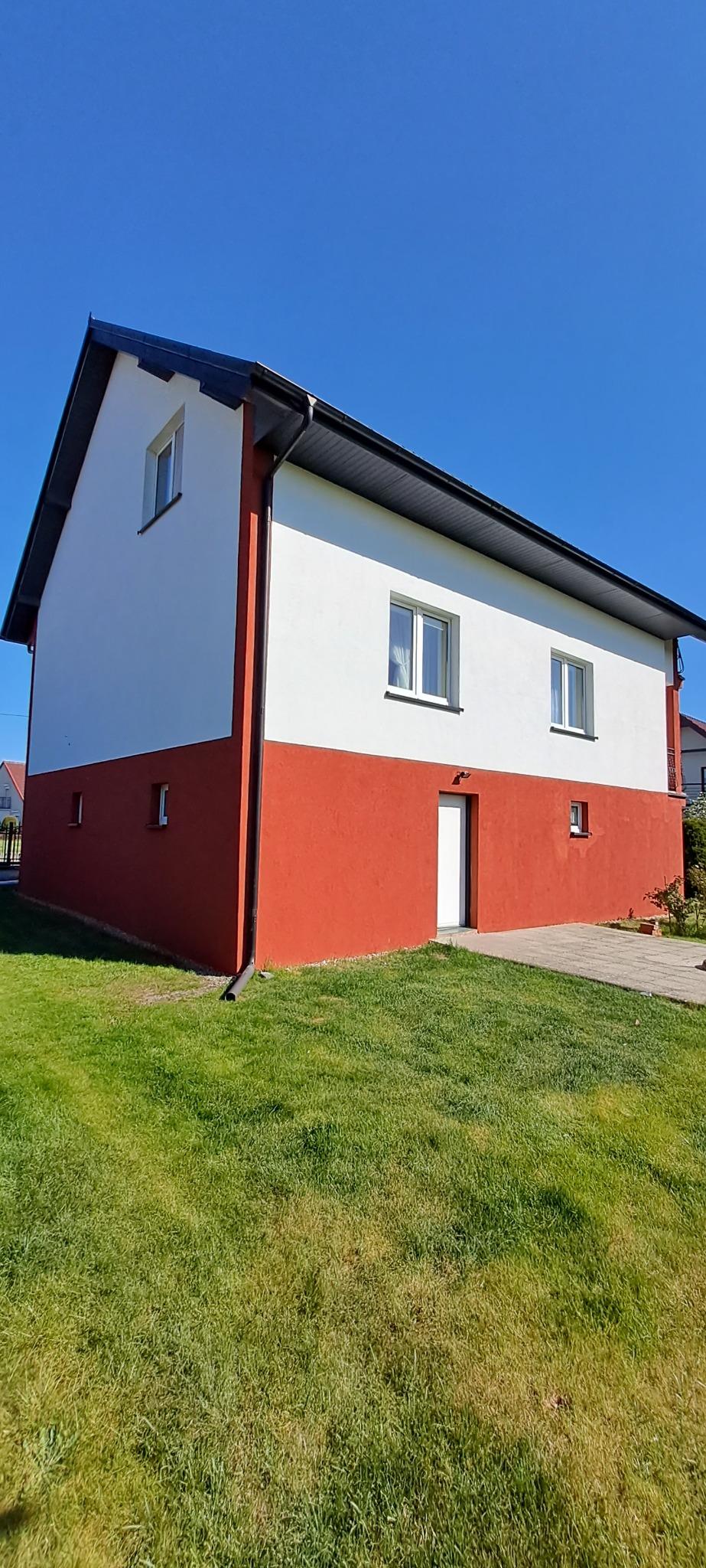 Dwukondygnacyjny dom z pomalowaną na biało górną częścią i na czerwono dolną, z czarnym dachem i rynnami, widok z zewnątrz na tle błękitnego nieba i zielonego trawnika.