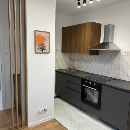 Furman-bud - Nowoczesna kuchnia w odcieniach szarości i drewna. Uwagę przykuwają podłużne płytki nad blatem oraz drewniana szafka wisząca. Na ścianie obrazek z drzewem i słońcem.