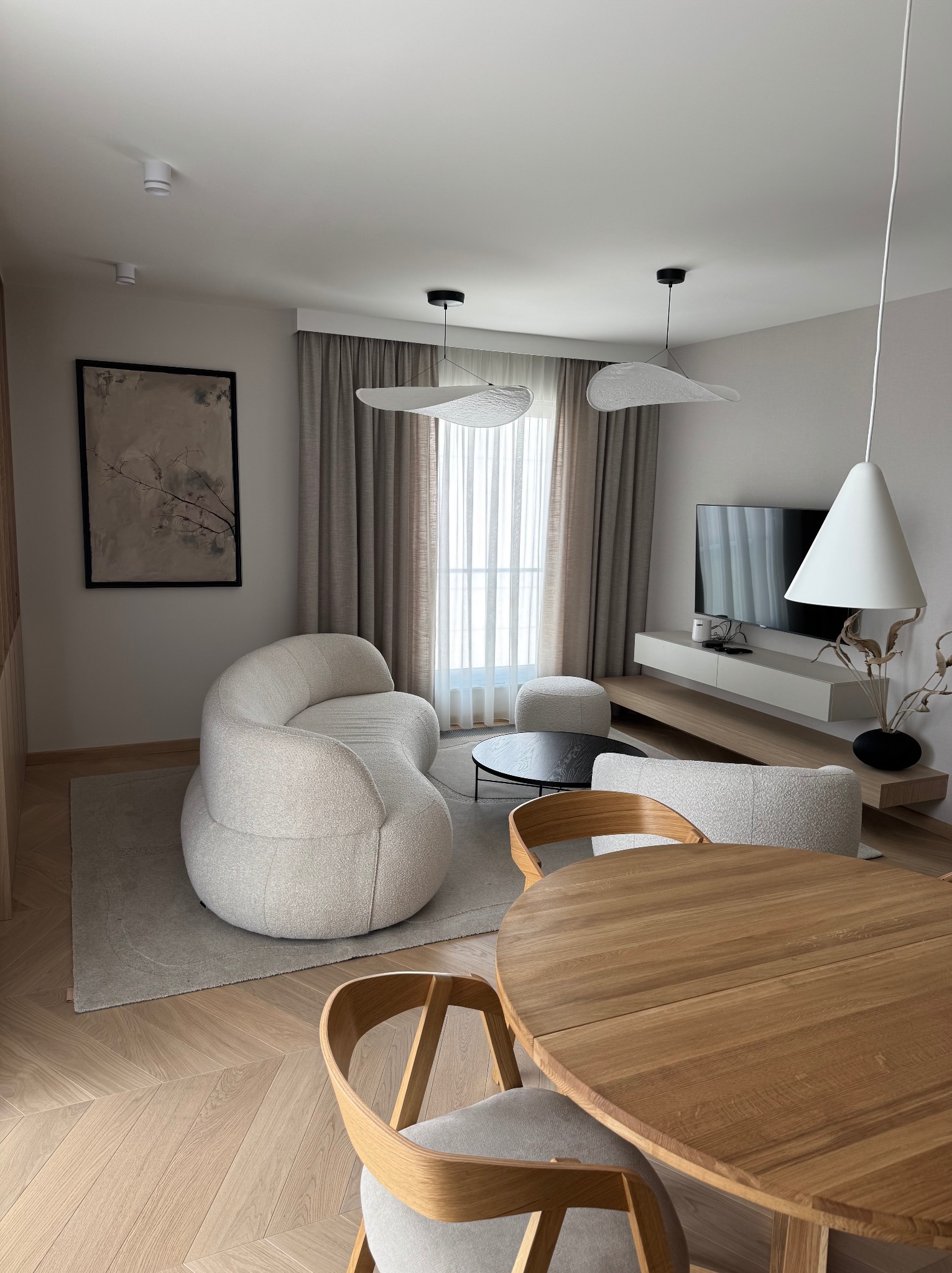 Stylowe, neutralne wnętrze: drewniany stół z krzesłami, sofa o opływowym kształcie, dywan, telewizor na szafce RTV, lampy wiszące. Parkiet w jodełkę, stonowana kolorystyka.