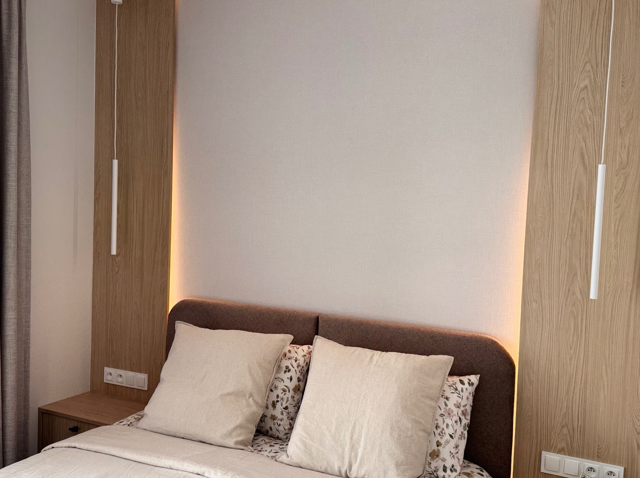 Nowoczesna sypialnia z oświetleniem LED w zagłówku, drewniane panele ścienne, minimalistyczne lampy wiszące i tekstylia w stonowanych barwach.