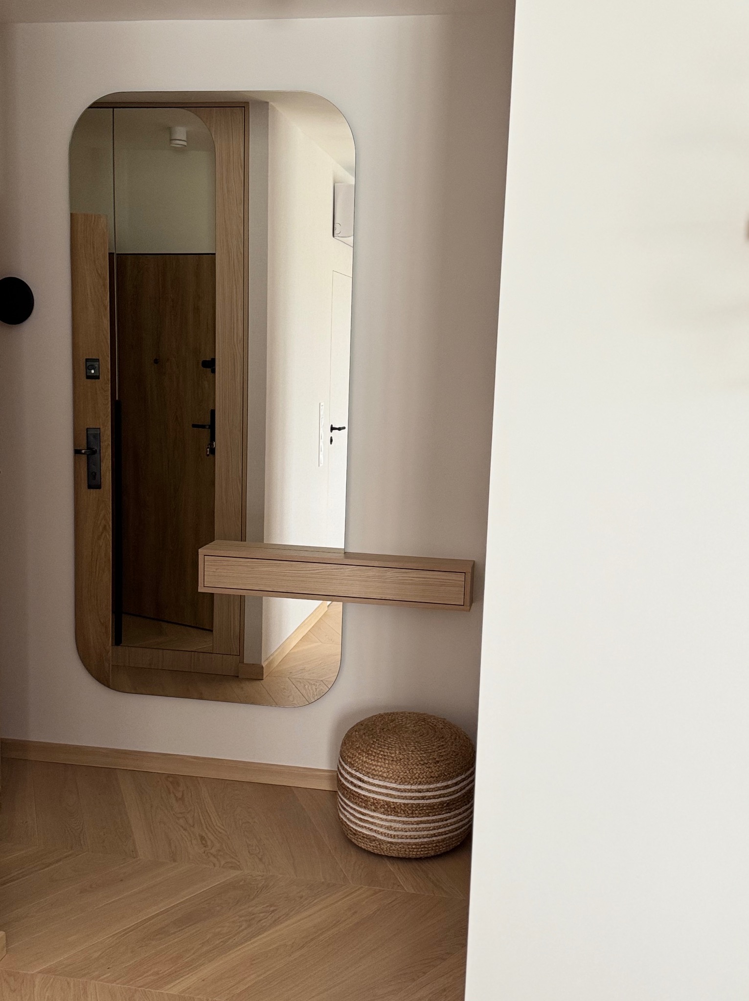 Nowoczesne lustro z półką w przedpokoju, odbijające drewniane drzwi i fragment wnętrza. Na podłodze parkiet w jodełkę, obok pufa ze sznurka.