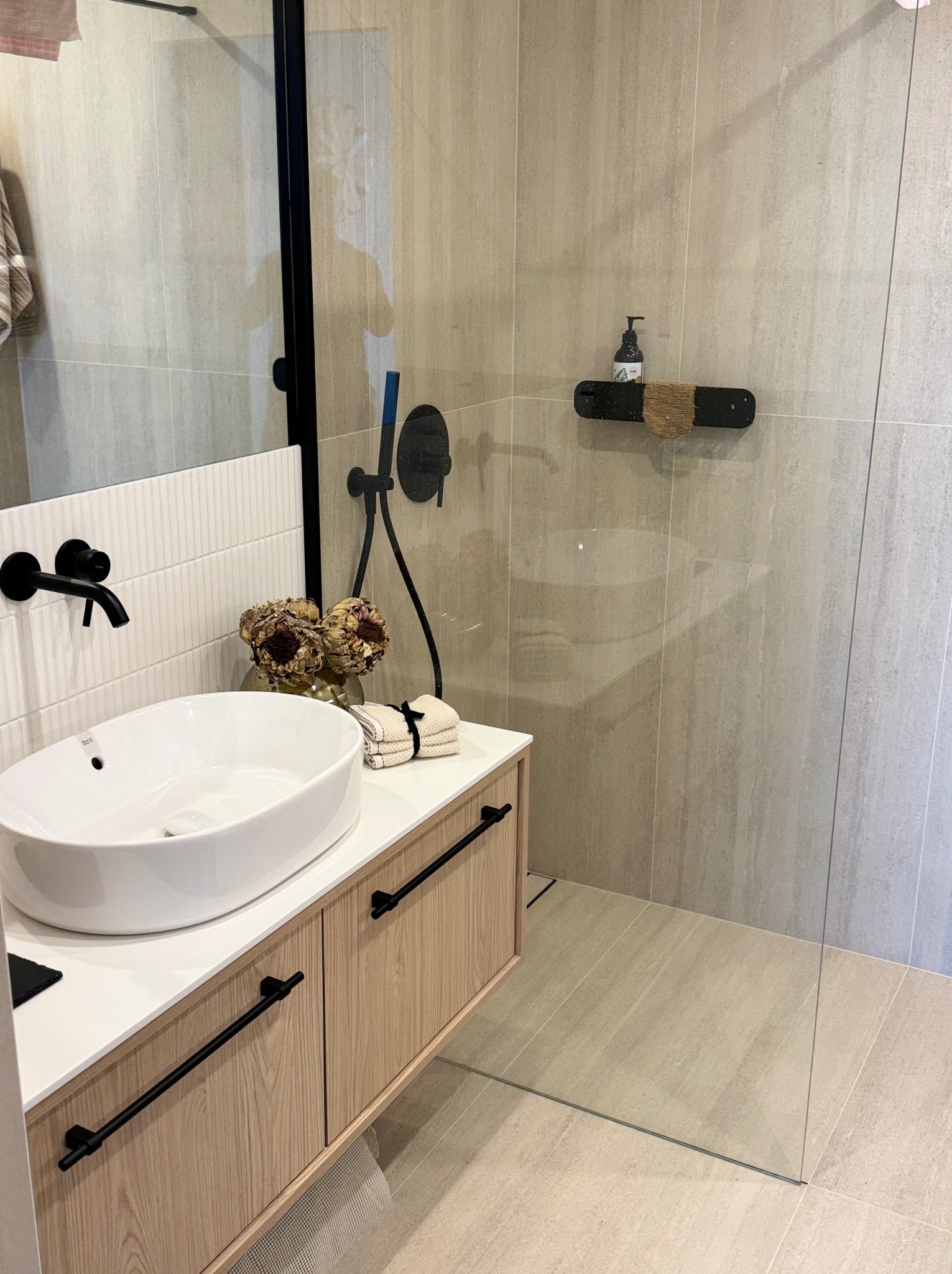 Nowoczesna aranżacja łazienki w Gdańsku: owalna umywalka na szafce z drewnianym fornirem, czarna armatura i kabina prysznicowa ze szkła, beżowe płytki.