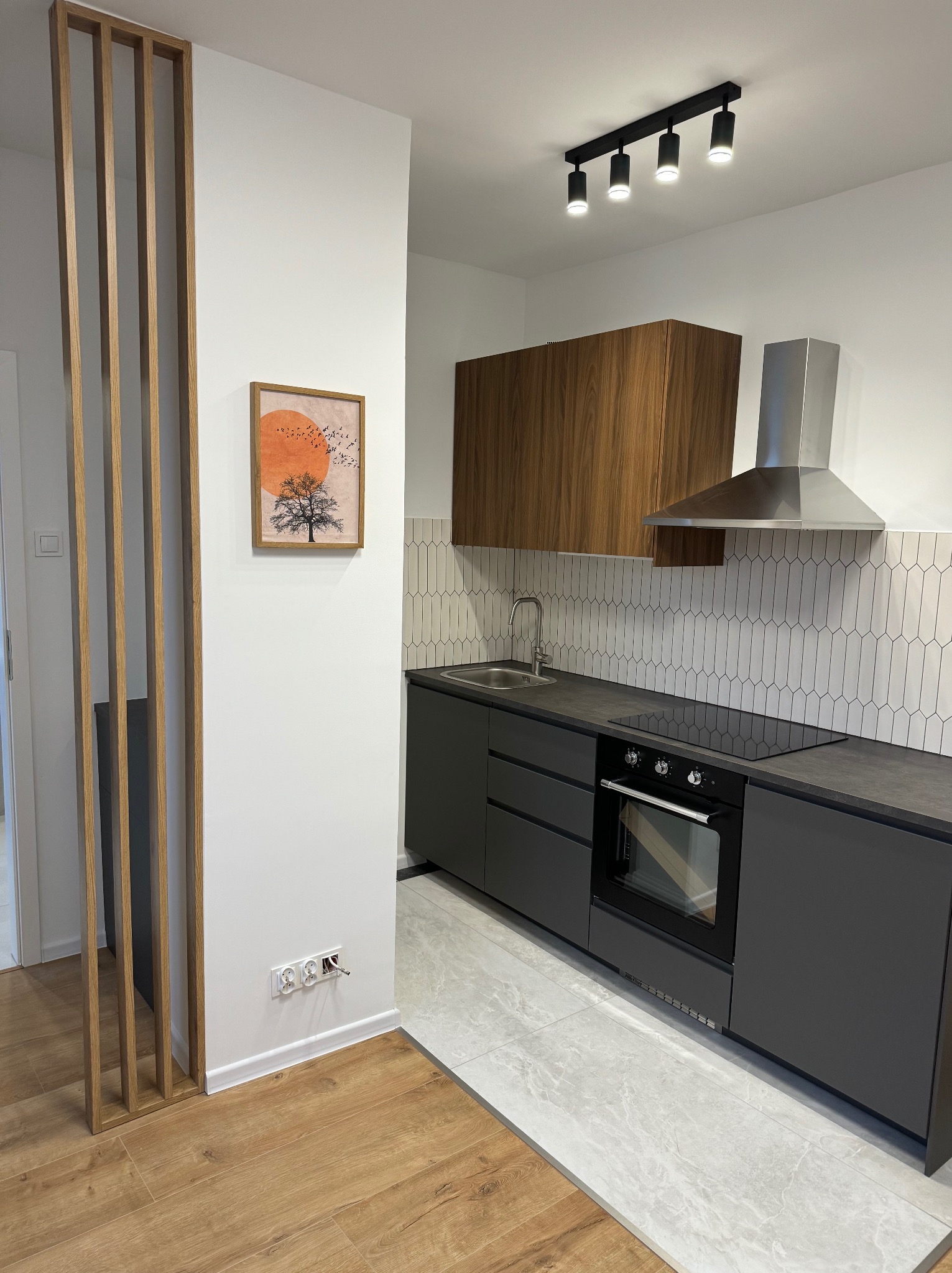 Nowoczesna kuchnia w odcieniach szarości i drewna. Uwagę przykuwają podłużne płytki nad blatem oraz drewniana szafka wisząca. Na ścianie obrazek z drzewem i słońcem.