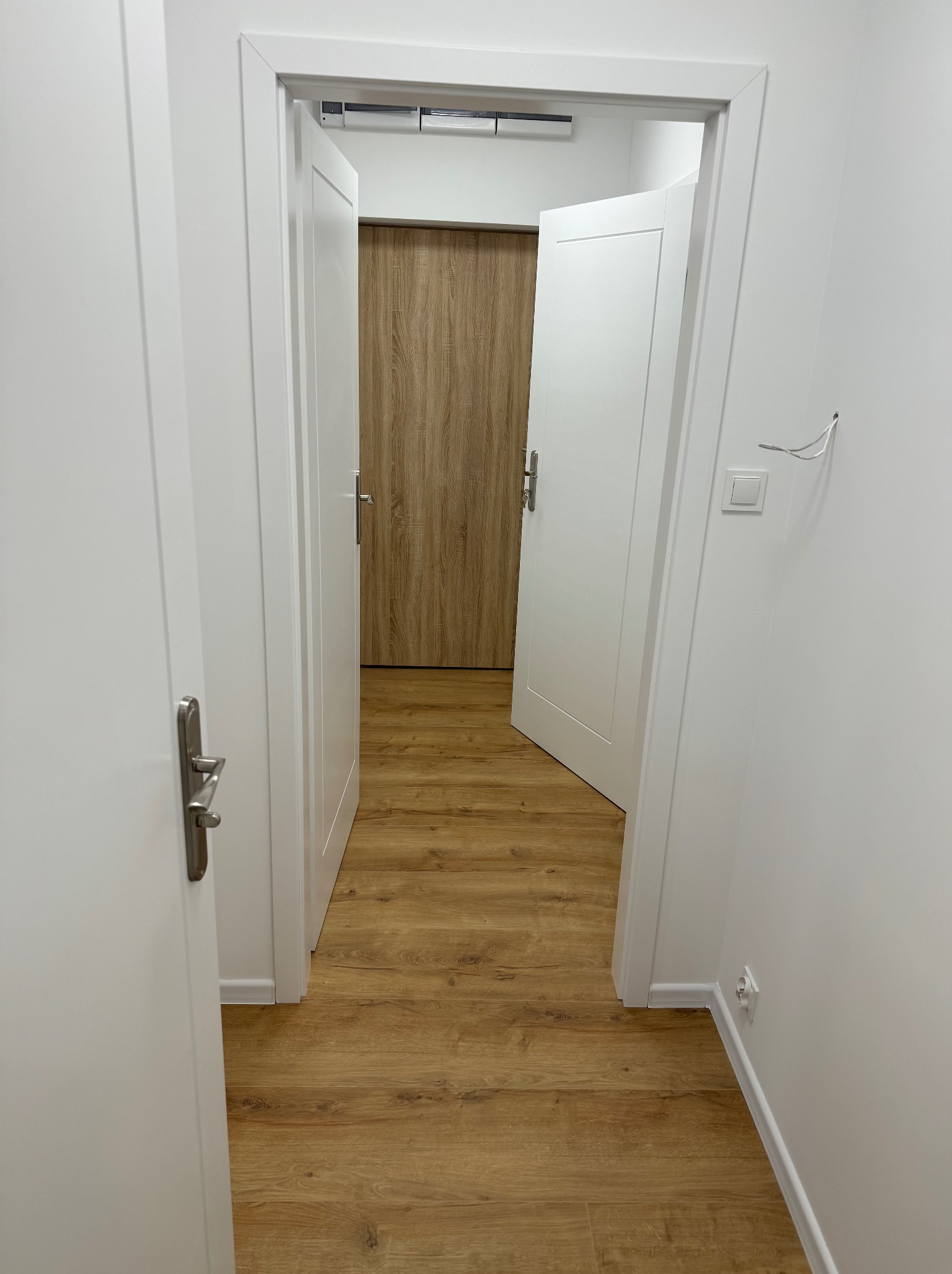 Wykończony korytarz z drewnianą podłogą, białymi ścianami i dwiema parami białych drzwi. Jedne drzwi są otwarte, ukazując dalszą część korytarza.