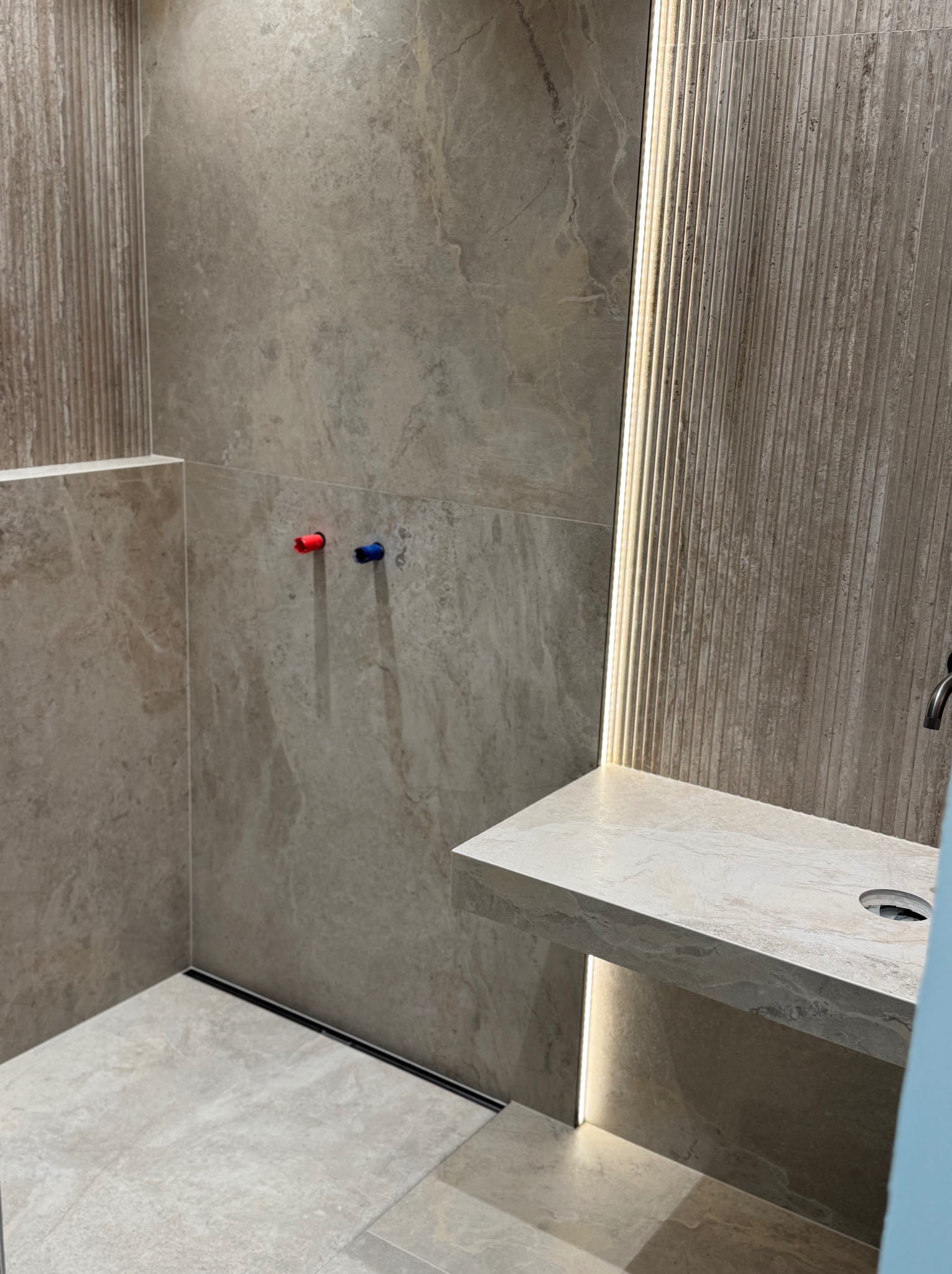 Beżowa łazienka z płytkami imitującymi kamień, z podświetleniem LED i minimalistycznym odpływem liniowym. Widoczne przyłącza wody z kolorowymi oznaczeniami.