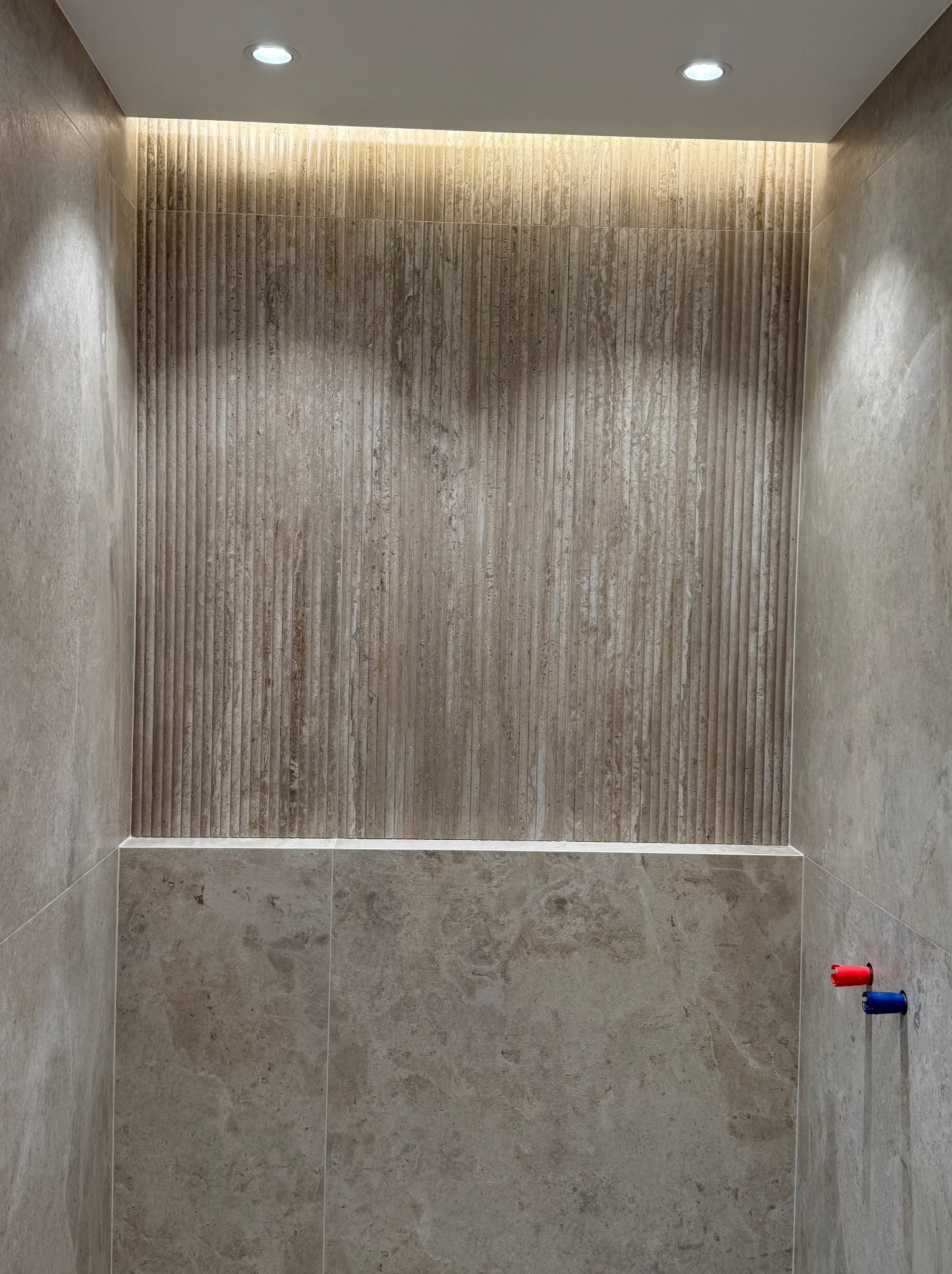Wykończona ściana w łazience z dekoracyjnymi, pionowo żłobionymi płytkami, podświetlona listwą LED. Widoczne zaślepki na rury wodne.
