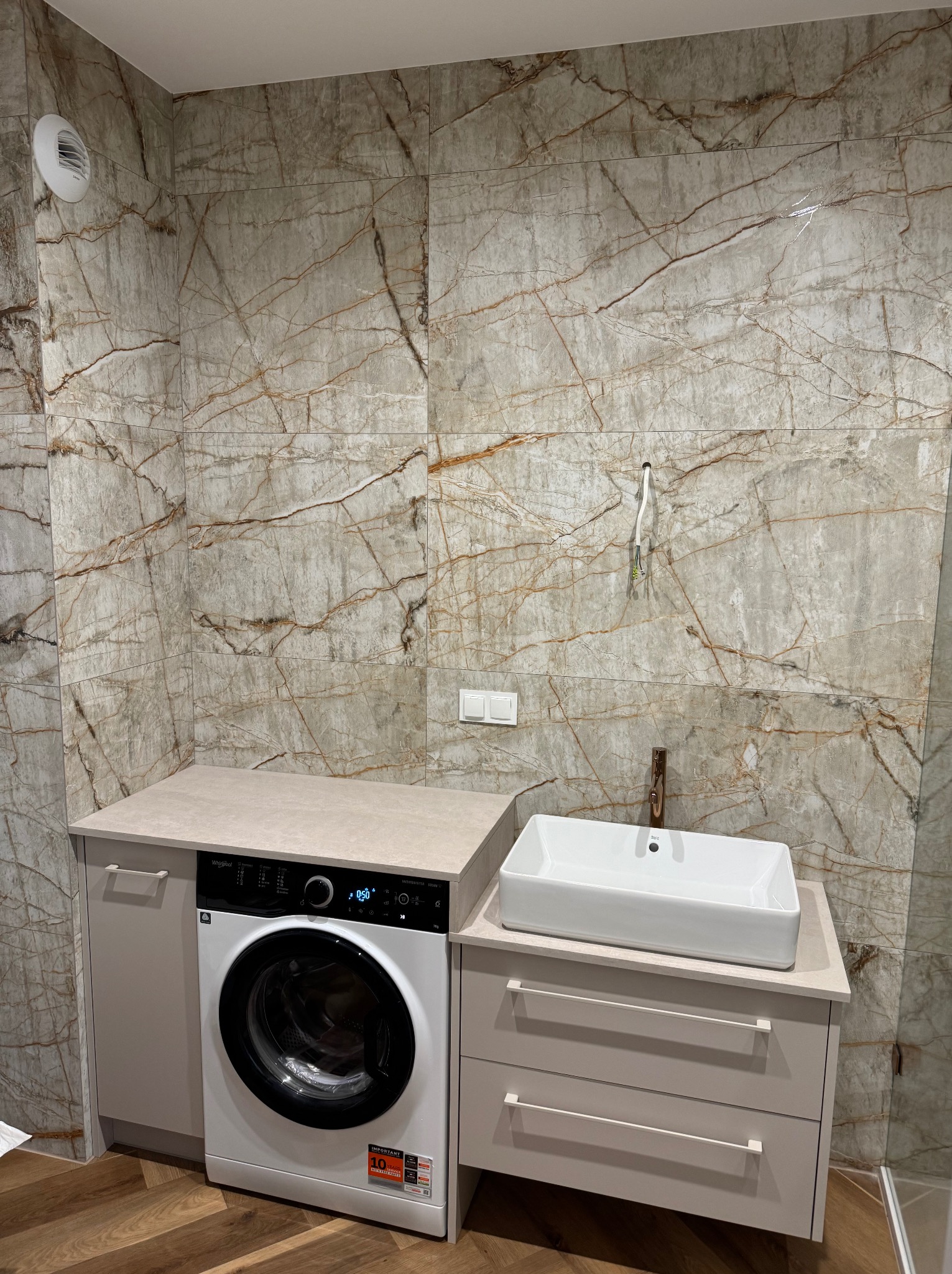Wykończona łazienka z marmurowymi płytkami, umywalką nablatową i pralką pod blatem. Widoczne przewody elektryczne przygotowane do podłączenia oświetlenia.