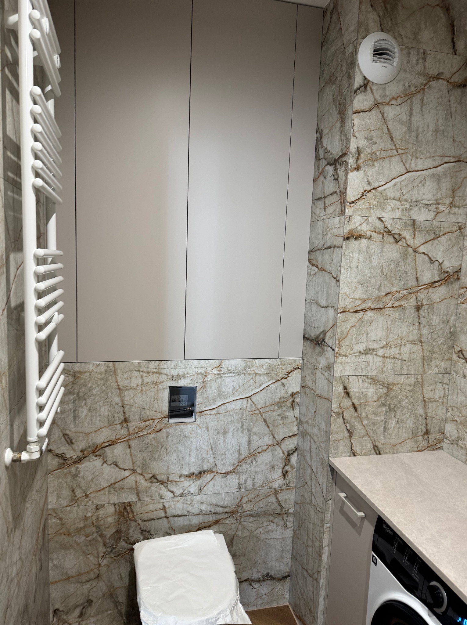 Nowoczesna łazienka z marmurowymi płytkami, grzejnikiem drabinkowym, zabudową meblową i pralką. Uwagę przyciąga wykończenie w odcieniach beżu i brązu oraz minimalistyczny design.