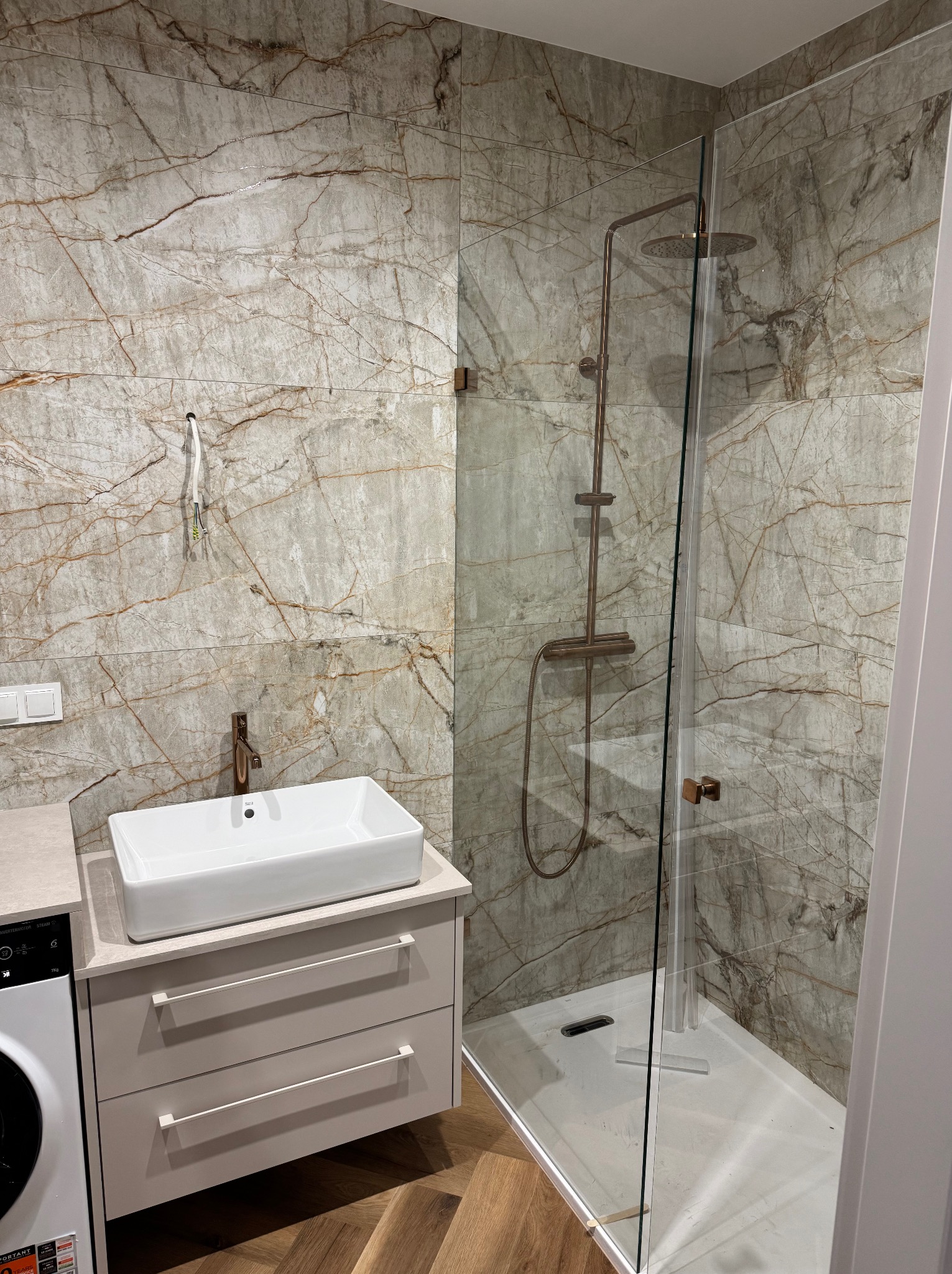 Nowoczesna łazienka z kabiną prysznicową typu walk-in, marmurowe płytki ścienne z brązowymi żyłami, biała umywalka nablatowa i armatura w kolorze różowego złota.