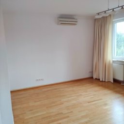 LT Łukasz Tabor - Pusty pokój z parkietem, klimatyzacją na ścianie, zasłoniętym oknem i lampą sufitową z niebieskim elementem.