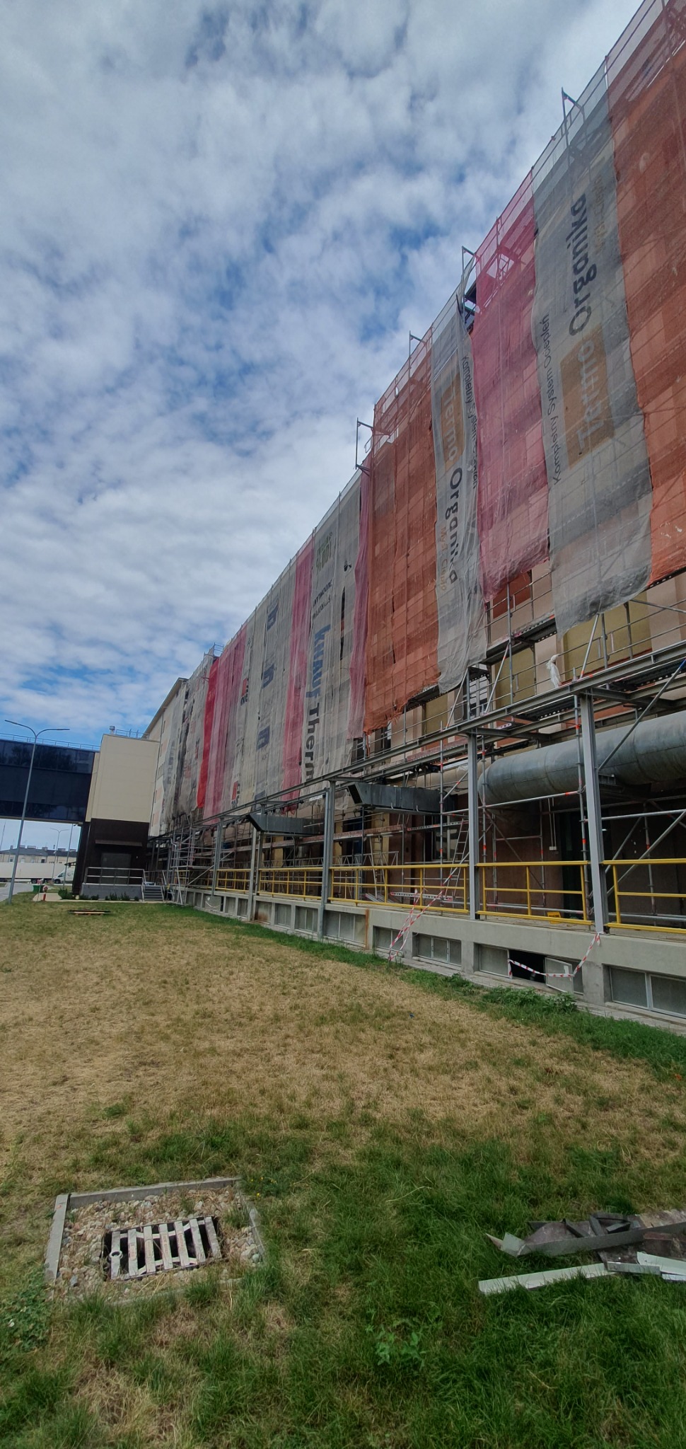 Wysoki budynek przemysłowy w trakcie prac elewacyjnych, osłonięty siatką z reklamami, widoczne rusztowania i rury, trawnik z kratką ściekową na pierwszym planie.