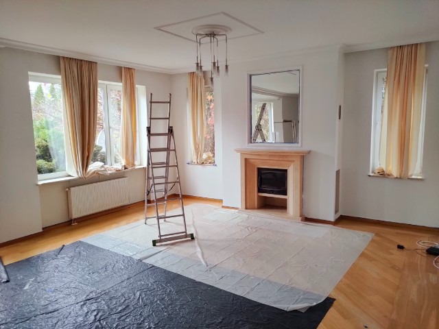 Pokój w trakcie malowania, zabezpieczona podłoga folią, drabina rozstawiona, ściany przygotowane do malowania, widoczny kominek z lustrem.