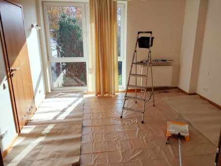 Pokój przygotowany do prac wykończeniowych: podłoga zabezpieczona folią ochronną, drabina malarska z kuwetą stoi przy grzejniku, widoczny wałek malarski.