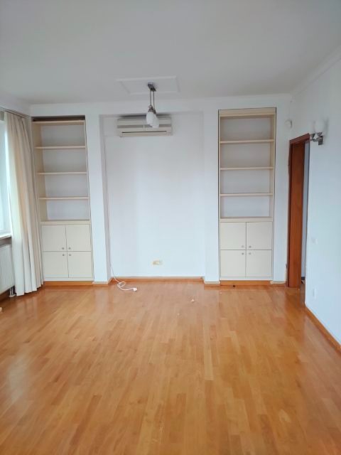 Pusty pokój z drewnianą podłogą, wbudowanymi regałami z szafkami po bokach, klimatyzatorem pod sufitem i białymi ścianami, gotowy do dalszych prac wykończeniowych.