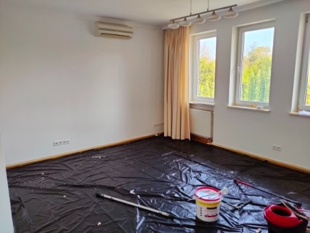 Pokój przygotowany do prac wykończeniowych: podłoga zabezpieczona czarną folią, widoczne narzędzia malarskie i wiadro z farbą, ściany pomalowane na biało, okna z kremowymi zasłonami.