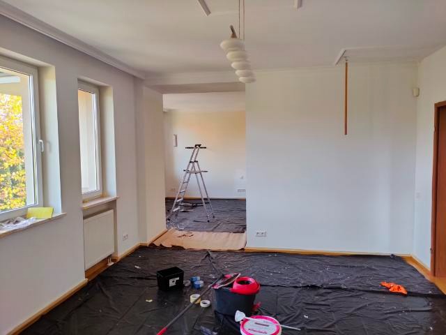 Przygotowanie do prac malarskich: pokój zabezpieczony folią malarską, widoczne narzędzia malarskie, drabina aluminiowa w tle, świeżo pomalowane ściany na biało.