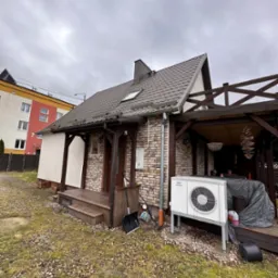 Zewnętrzna jednostka klimatyzacji typu split zainstalowana przy ceglanym domu z drewnianym zadaszeniem tarasu. Na pierwszym planie widoczny trawnik z kamieniami, w tle budynek mieszkalny.