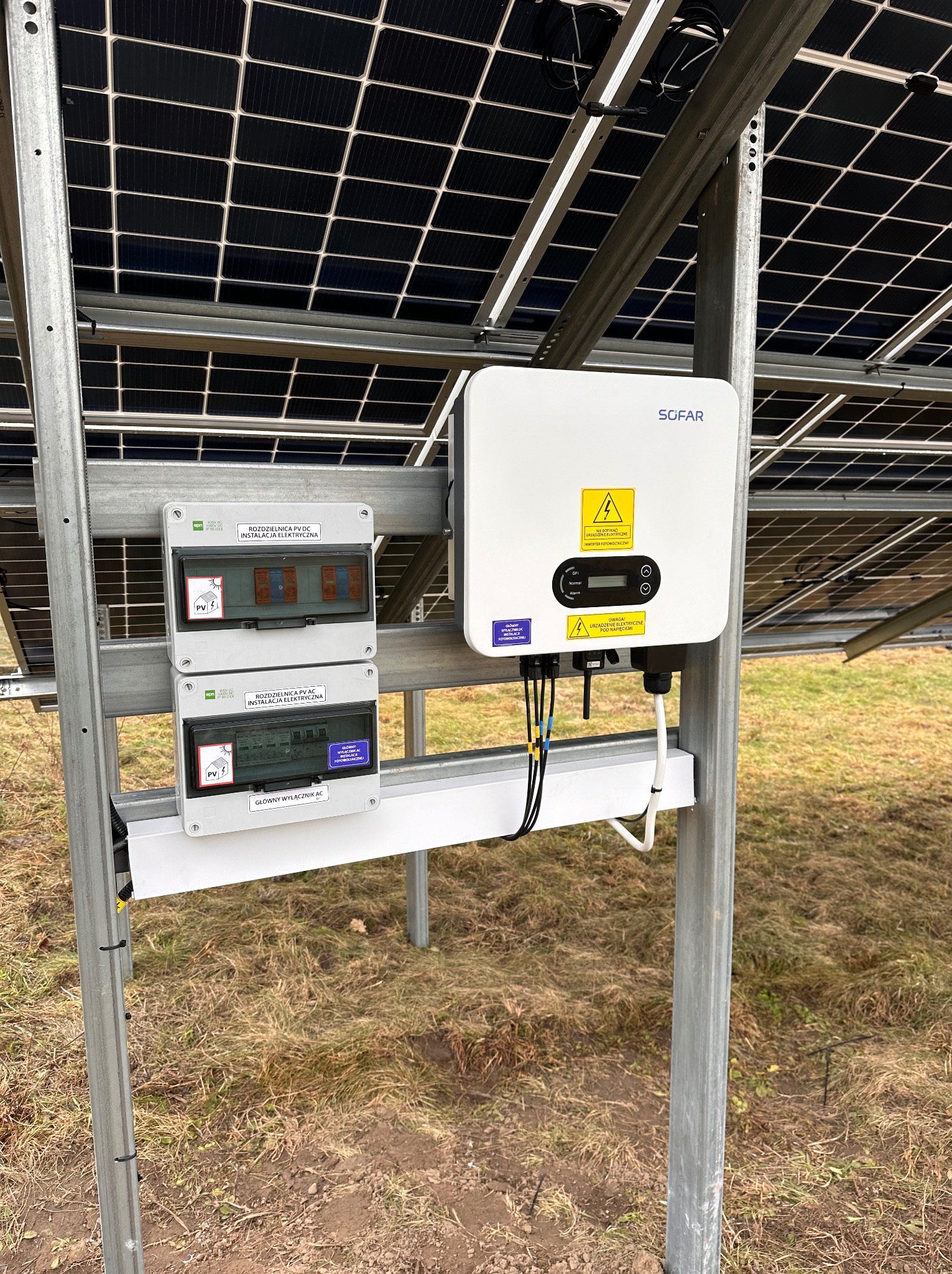 Instalacja fotowoltaiczna: widok na inwerter SOFAR, rozdzielnice AC i DC oraz moduły fotowoltaiczne na metalowej konstrukcji, na tle trawy.