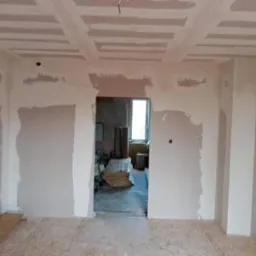 Wnętrze w trakcie prac wykończeniowych: ściany częściowo pokryte gładzią, sufit z widoczną konstrukcją pod płyty kartonowo-gipsowe, podłoga z płyt OSB, widok na pomieszczenie obok przez otw&oacute;r...