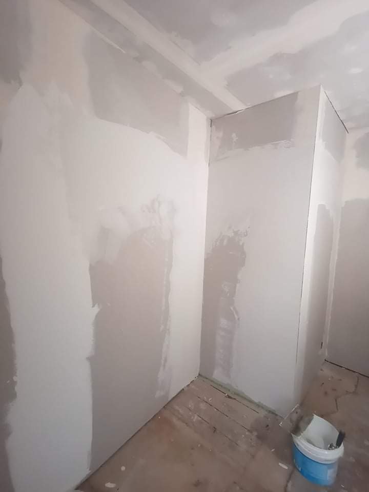 Pokój w trakcie prac wykończeniowych: ściany z nałożoną warstwą gładzi gipsowej, widoczna surowa płyta karton-gips na suficie, podłoga drewniana częściowo zakryta, wiadro z narzędziami w rogu...