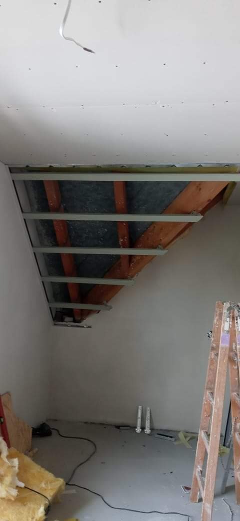 Skośny sufit poddasza w trakcie montażu stelażu z profili metalowych pod płyty gipsowo-kartonowe, widoczne drewniane elementy konstrukcyjne dachu i częściowo ocieplenie, drabina oparta o ścianę.