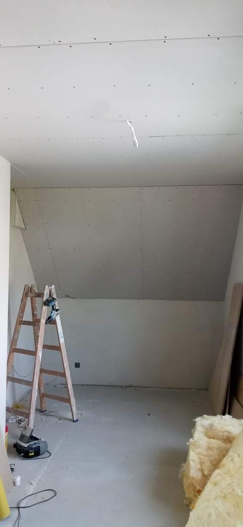 Poddasze w trakcie wykańczania płytami kartonowo-gipsowymi, widoczna drewniana drabina z wkrętarką, częściowo ocieplone wełną mineralną.