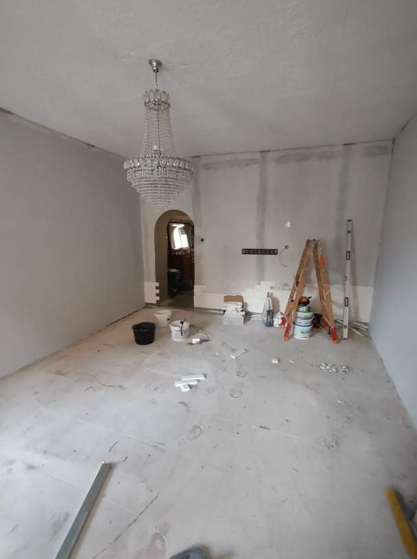 Pokój w trakcie prac wykończeniowych: ściany przygotowane pod malowanie, widoczne ślady szpachlowania, drabina oparta o ścianę, wiadro z farbą, luksusowy kryształowy żyrandol nadaje kontrastu...