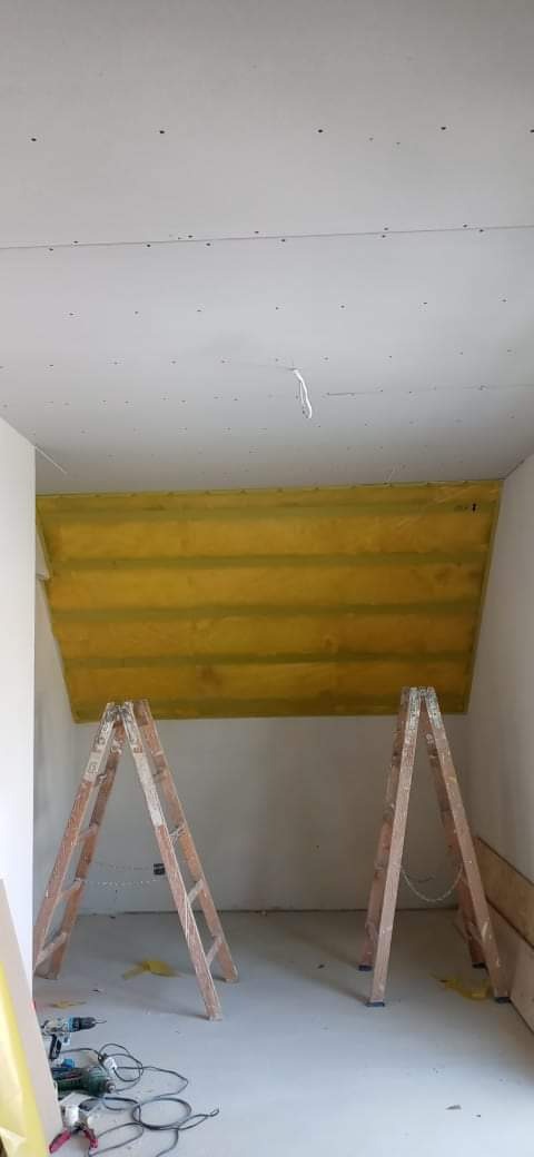 Skośny sufit poddasza z izolacją z wełny mineralnej, widoczne drewniane legary, częściowo zakryte płytą karton-gips, dwie drewniane drabiny, narzędzia na podłodze.