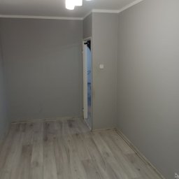Doradca L.J.Builders - Szare, świeżo pomalowane wnętrze pokoju z jasną podłogą imitującą drewno. Widoczna lampa sufitowa i fragment drzwi.