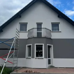 Elewacja domu po malowaniu, z szarym pasem na środku. Widoczne rusztowanie, cegły na chodniku i balkon z czarną, ozdobną balustradą.