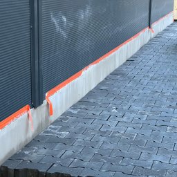 Doradca L.J.Builders - Szara elewacja budynku zabezpieczona pomarańczową taśmą malarską, z widoczną szarą kostką brukową i częścią sylwetki pracownika w prawym górnym rogu.