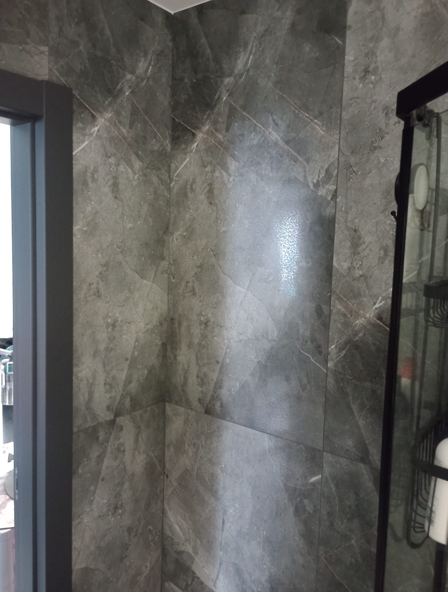 Wykończona łazienka z szarymi, marmurkowymi płytkami na ścianach i kabiną prysznicową z czarnymi profilami. Widoczny fragment ościeżnicy drzwi.