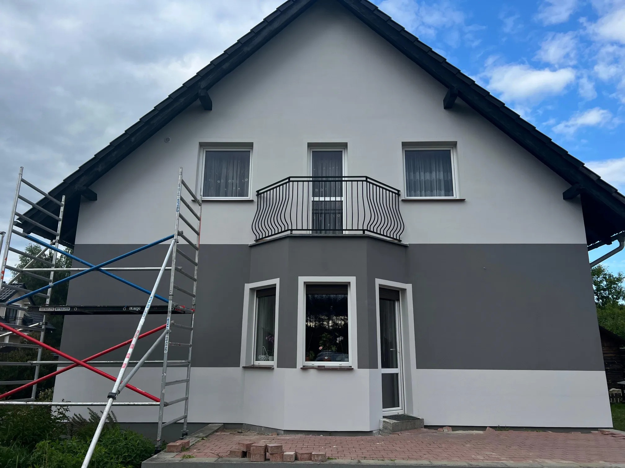 Elewacja domu po malowaniu, z szarym pasem na środku. Widoczne rusztowanie, cegły na chodniku i balkon z czarną, ozdobną balustradą.