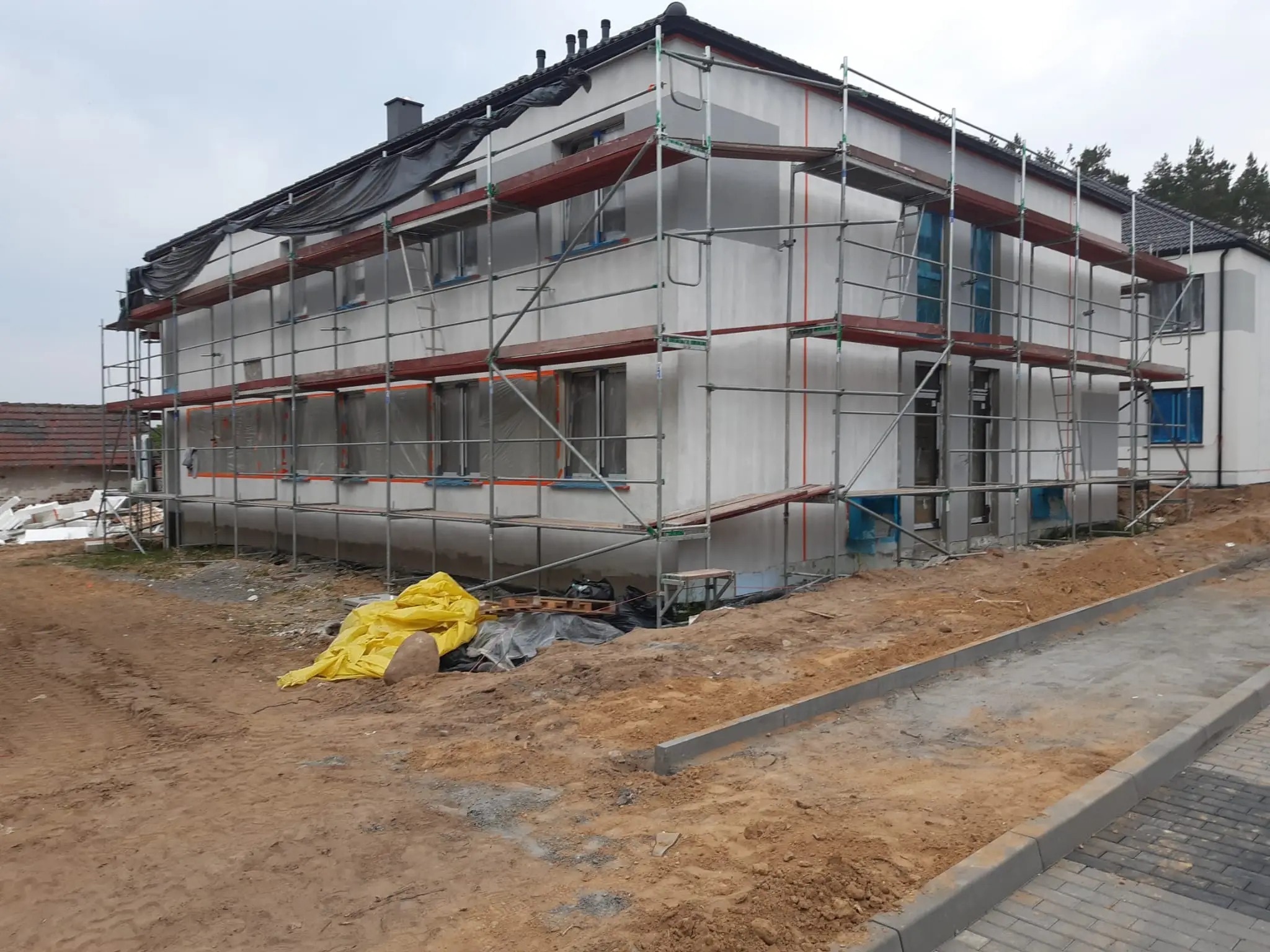 Dwukondygnacyjny dom w trakcie prac elewacyjnych, otoczony rusztowaniem i plandekami, z widocznymi przygotowaniami do dalszych prac wykończeniowych.