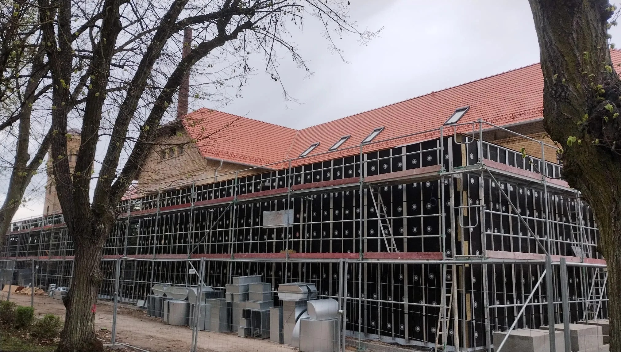 Budynek w trakcie ocieplania, widoczne rusztowania i czarne płyty izolacyjne. Na pierwszym planie metalowe elementy wentylacyjne. Czerwony dach i drzewa w tle.