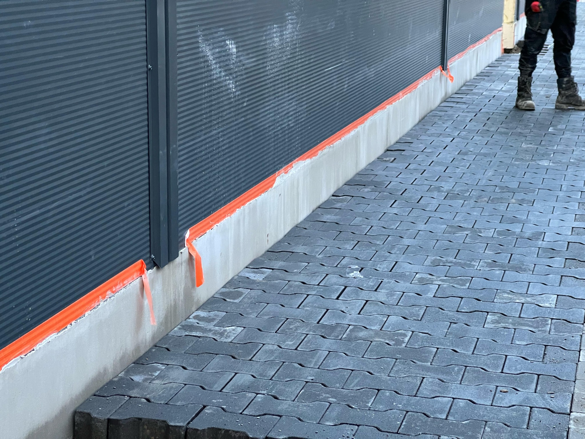 Szara elewacja budynku zabezpieczona pomarańczową taśmą malarską, z widoczną szarą kostką brukową i częścią sylwetki pracownika w prawym górnym rogu.