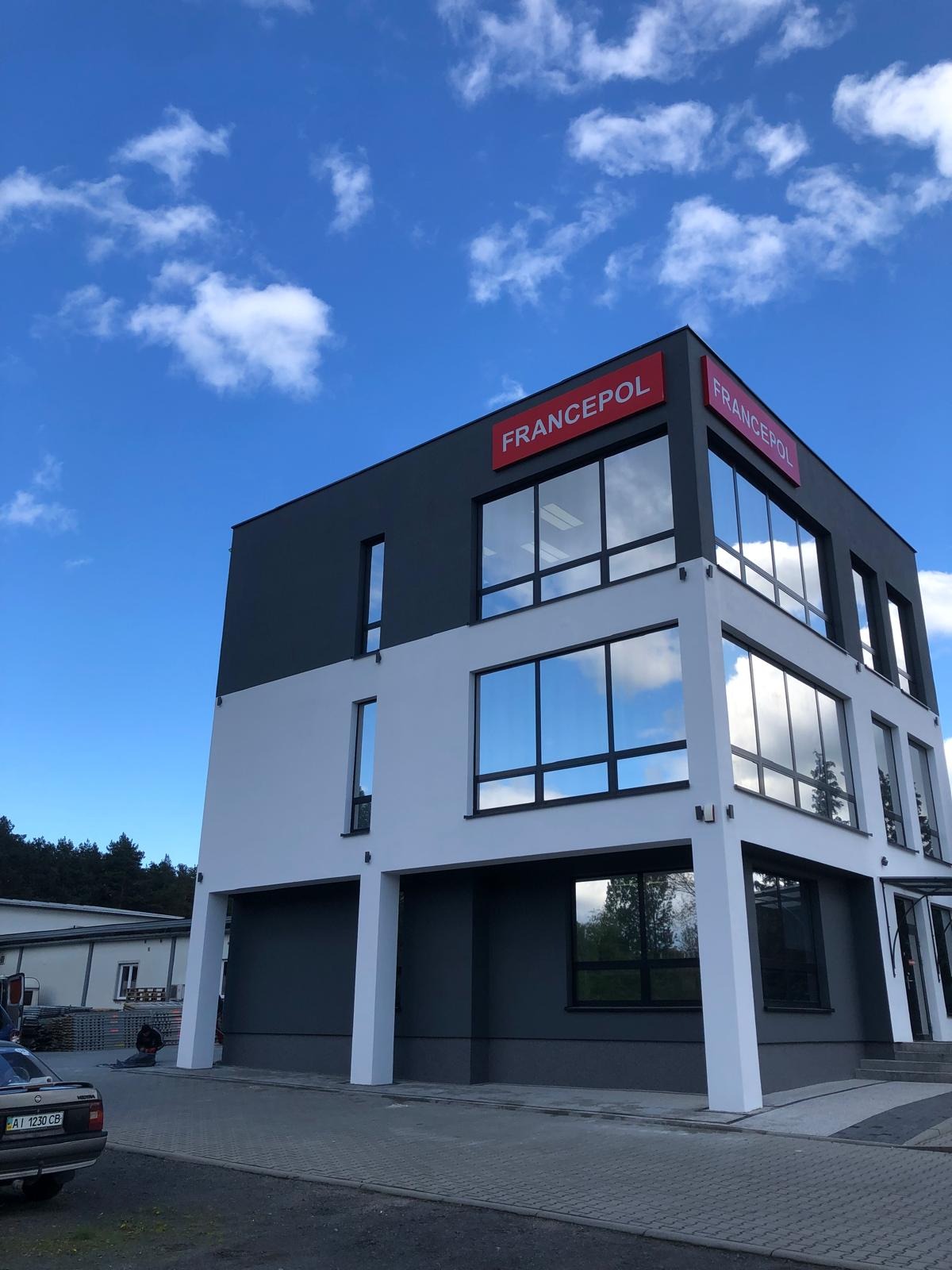 Dwukondygnacyjny budynek biurowy w nowoczesnej kolorystyce: białe i grafitowe ściany, duże okna z odbiciem nieba, czerwone logo na dachu.