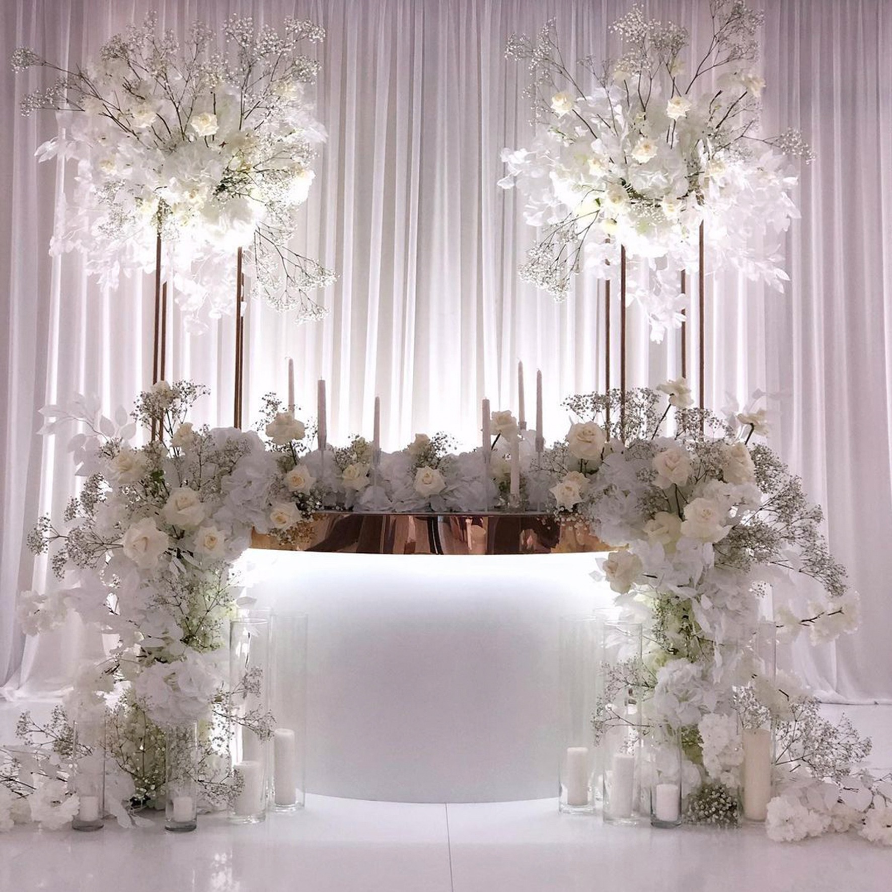 Dekoracja stołu weselnego w bieli: kompozycje kwiatowe z białych róż i gipsówki na złotych stojakach, świece w szklanych cylindrach, białe tło z draperii.