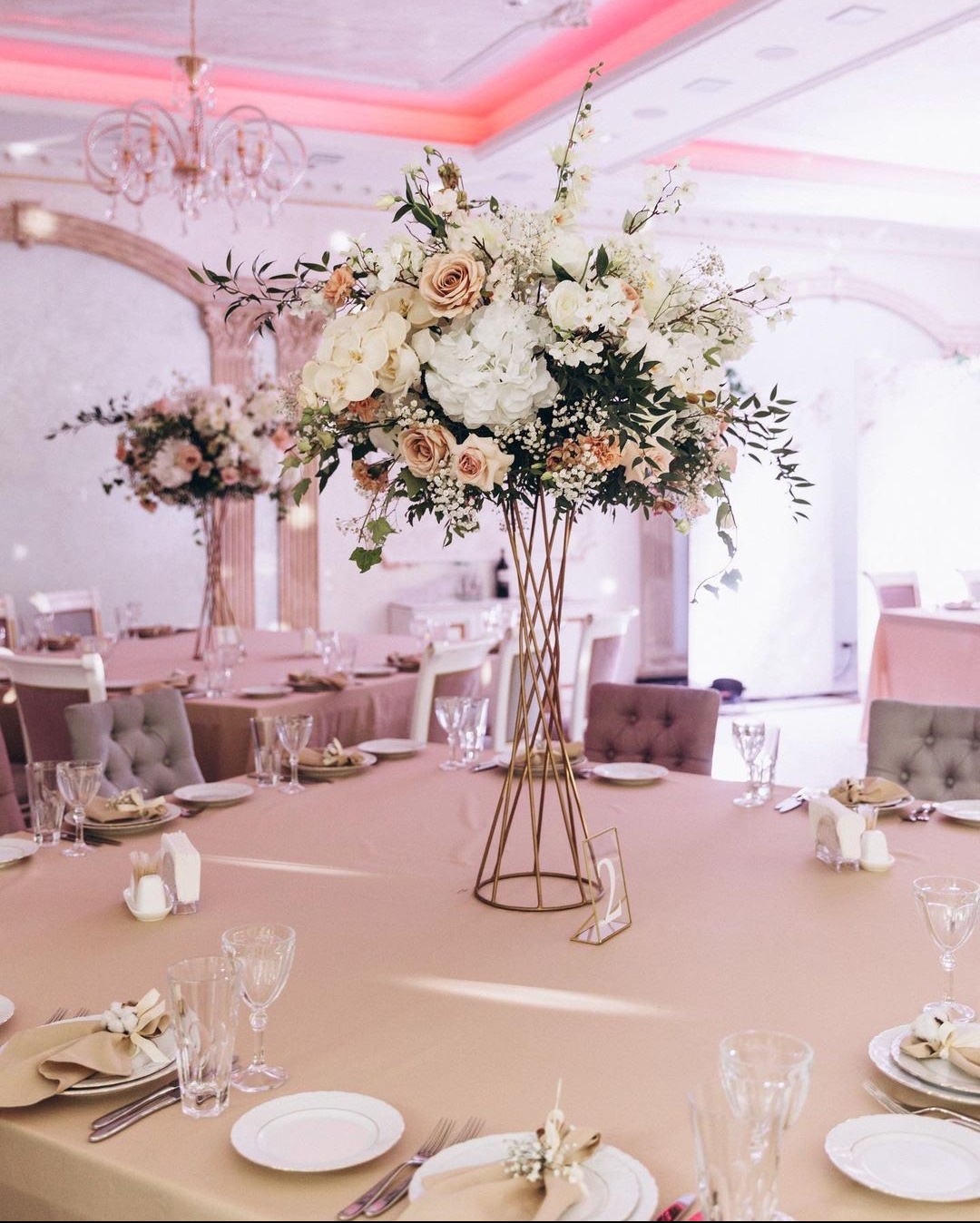 Elegancka kompozycja kwiatowa z białych i kremowych róż oraz zieleni w złotej, ażurowej podstawie na stole weselnym z numerem '2', nakrytym beżowym obrusem i zastawą.