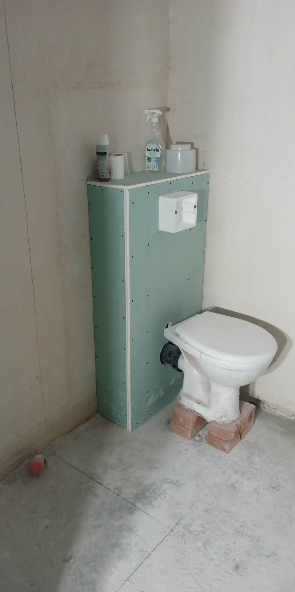 Niezakończona obudowa geberitu z płyt karton gips, tymczasowo zamontowana toaleta na cegłach, butelka Ludwik na obudowie, surowe ściany i podłoga w trakcie remontu łazienki.