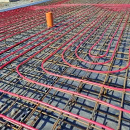 Czerwone rury ogrzewania podłogowego ułożone na stalowej siatce zbrojeniowej, widoczne elementy szalunku i wystające rury kanalizacyjne.