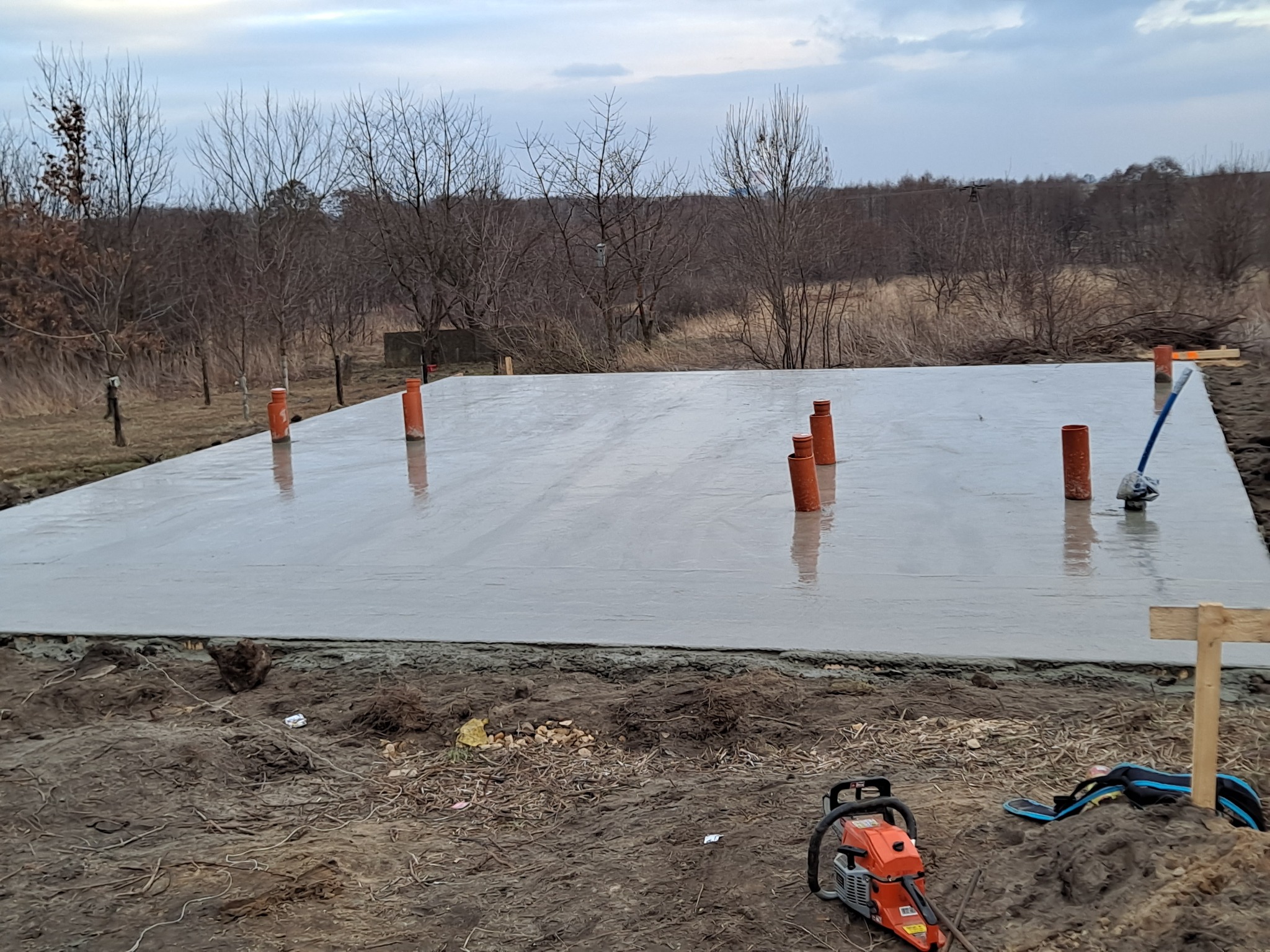 Wylana płyta fundamentowa z wystającymi rurami kanalizacyjnymi, na pierwszym planie ubijarka wibracyjna i torba robocza, w tle drzewa bez liści pod pochmurnym niebem.