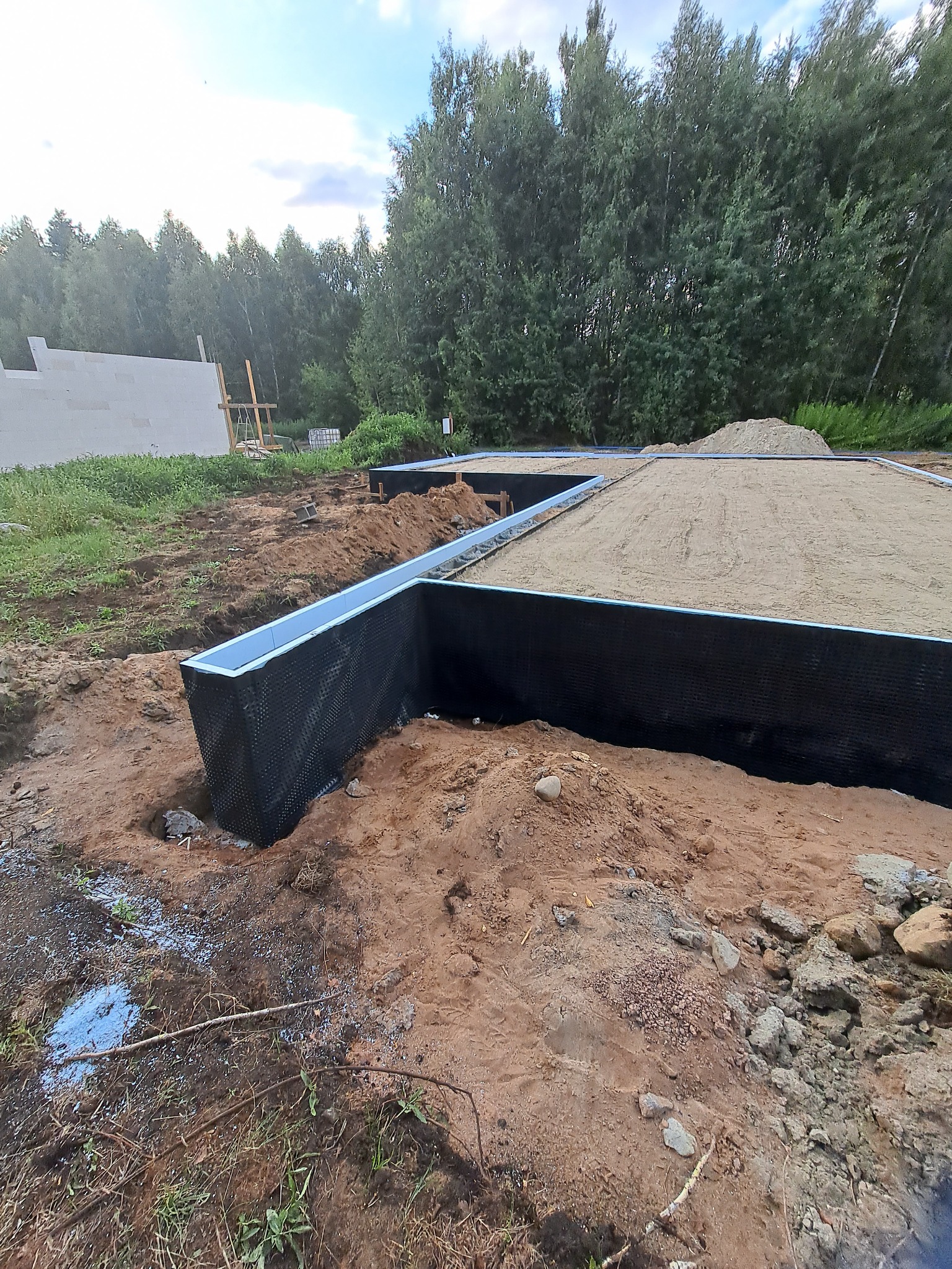 Przygotowany fundament pod budynek z izolacją pionową, zasypany piaskiem, widok na teren budowy z drzewami w tle.