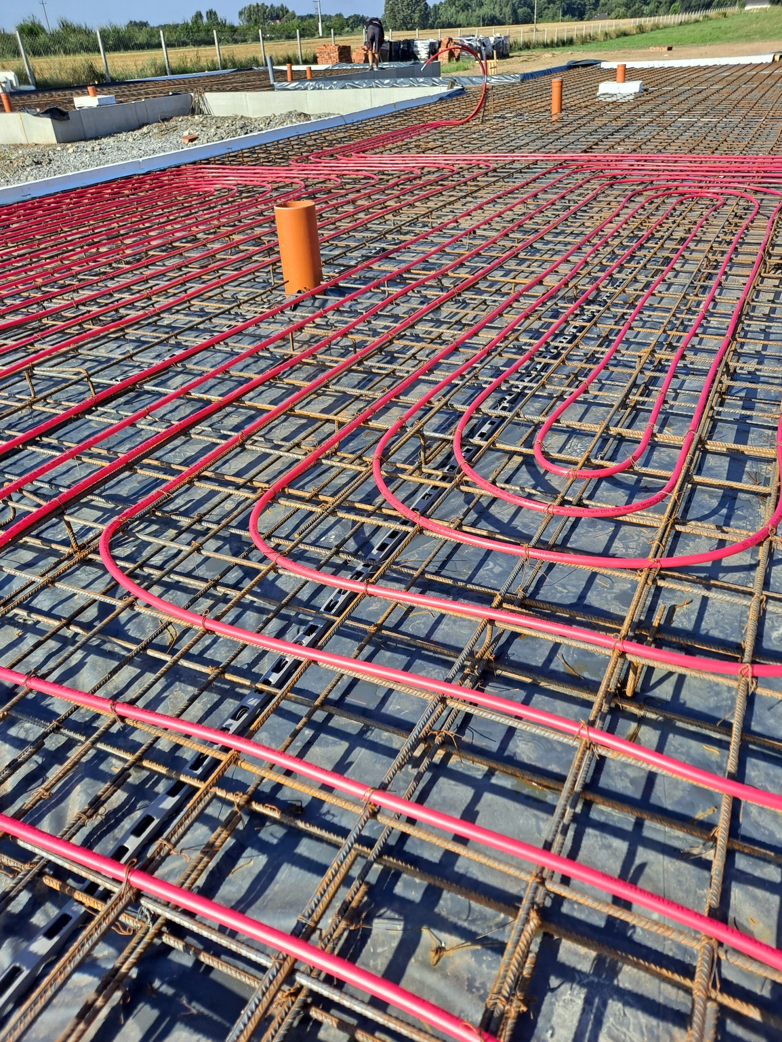 Czerwone rury ogrzewania podłogowego ułożone na stalowej siatce zbrojeniowej, widoczne elementy szalunku i wystające rury kanalizacyjne.