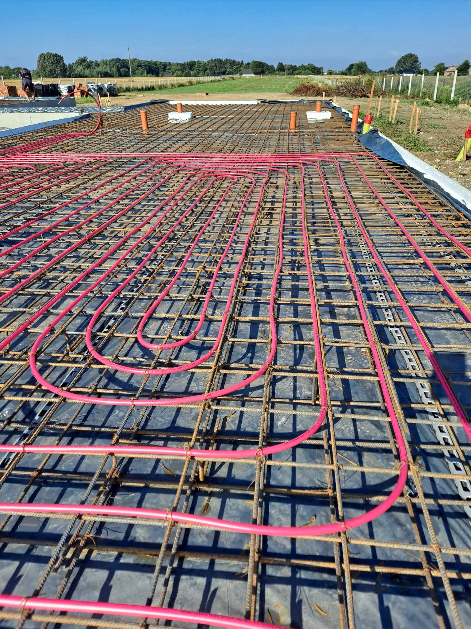 Zbrojenie fundamentu z rozłożonymi rurkami ogrzewania podłogowego w kolorze różowym, widoczne elementy szalunku i pracownicy w tle.